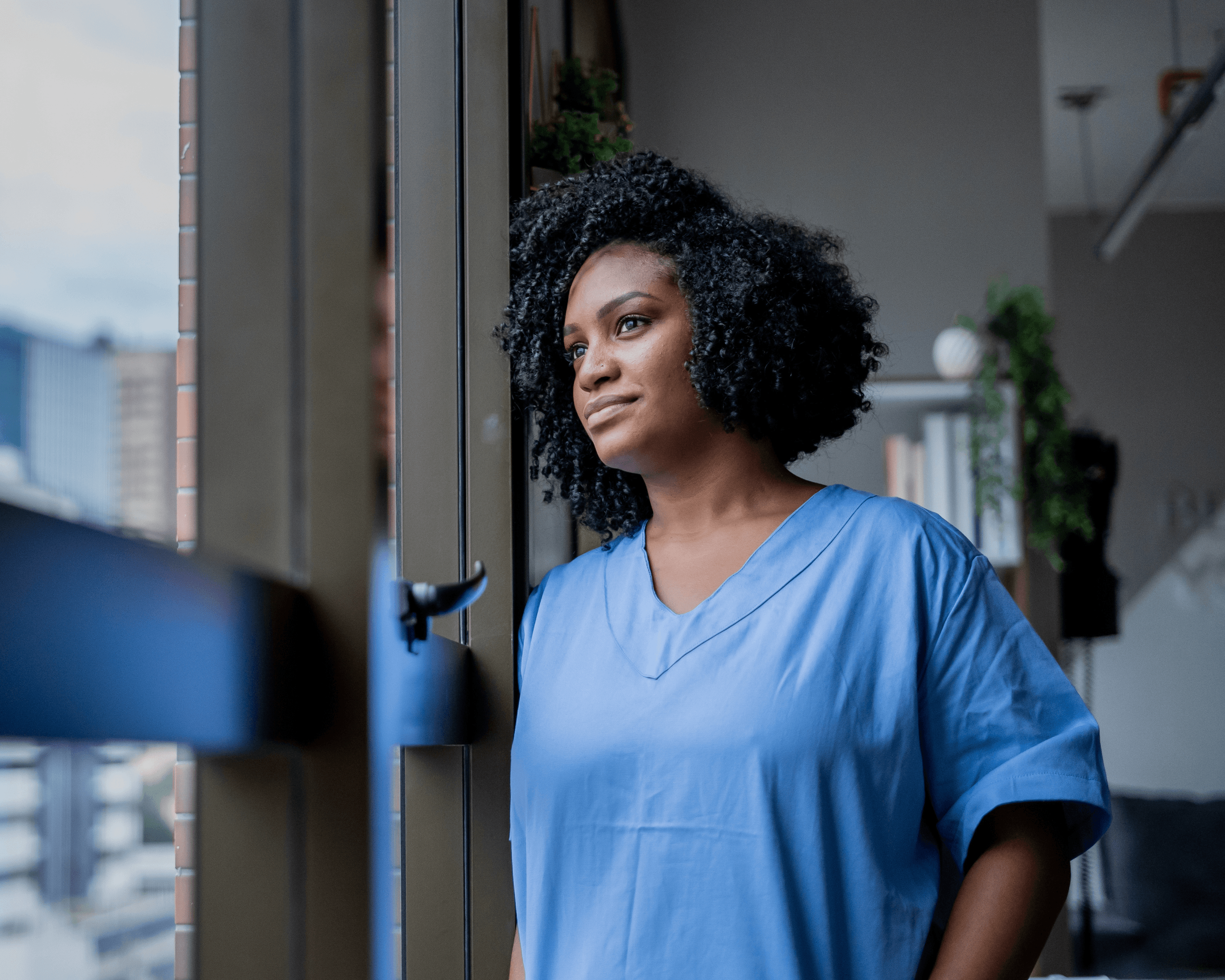 African woman wearing scrubs looks out a window 